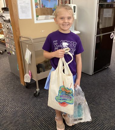 A young girl inside the Minneapolis Public Library holding up her “Color Our World” reading bag that she colored.