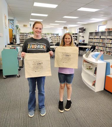 Two girls at the Clearwater Public Library holding up her “Color Our World” reading bags.