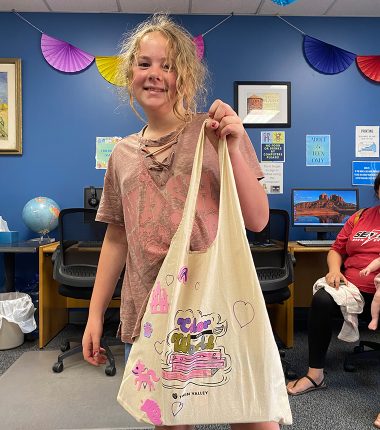 A young girl inside the Belle Plaine Public Library holding up her “Color Our World” reading bag that she colored.
