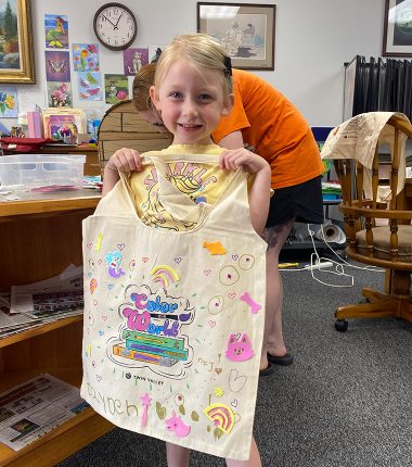A young girl inside the Belle Plaine Public Library holding up her “Color Our World” reading bag that she colored.