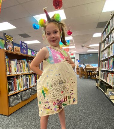 A young girl inside the Belle Plaine Public Library holding up her “Color Our World” reading bag that she colored.