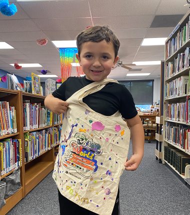 A young boy inside the Belle Plaine Public Library holding up his “Color Our World” reading bag that he colored.