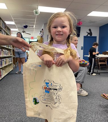 A young girl inside the Belle Plaine Public Library holding up her “Color Our World” reading bag that she colored.