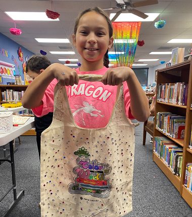 A young girl inside the Belle Plaine Public Library holding up her “Color Our World” reading bag that she colored.