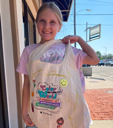 A young girl outside the Belle Plaine Public Library holding up her “Color Our World” reading bag that she colored.